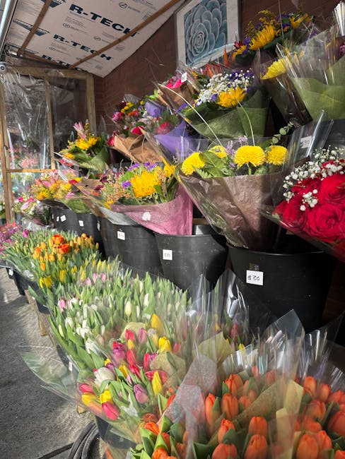 A display of fresh floral bouquets arranged in black pots and wrapped in colorful paper, featuring bright yellow chrysanthemums, purple and white daisies, red roses, pink and orange tulips, and mixed