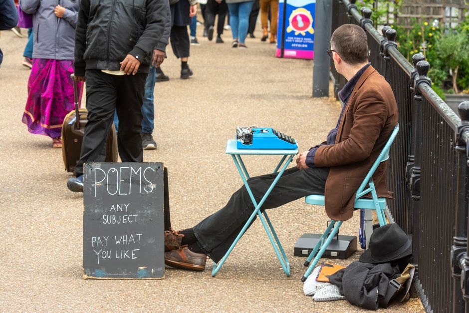 A man wearing glasses, a brown blazer, and black trousers is seated on a blue folding chair on a sidewalk, arranging flowers on a small blue table. The table holds a camera and a keyboard. Near his fe