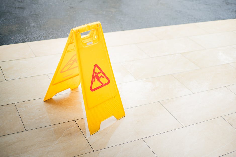 A yellow wet floor warning sign with red triangle symbols and an image of a person slipping is placed on a beige tiled floor near the edge of a polished surface, likely in a public or commercial indoo
